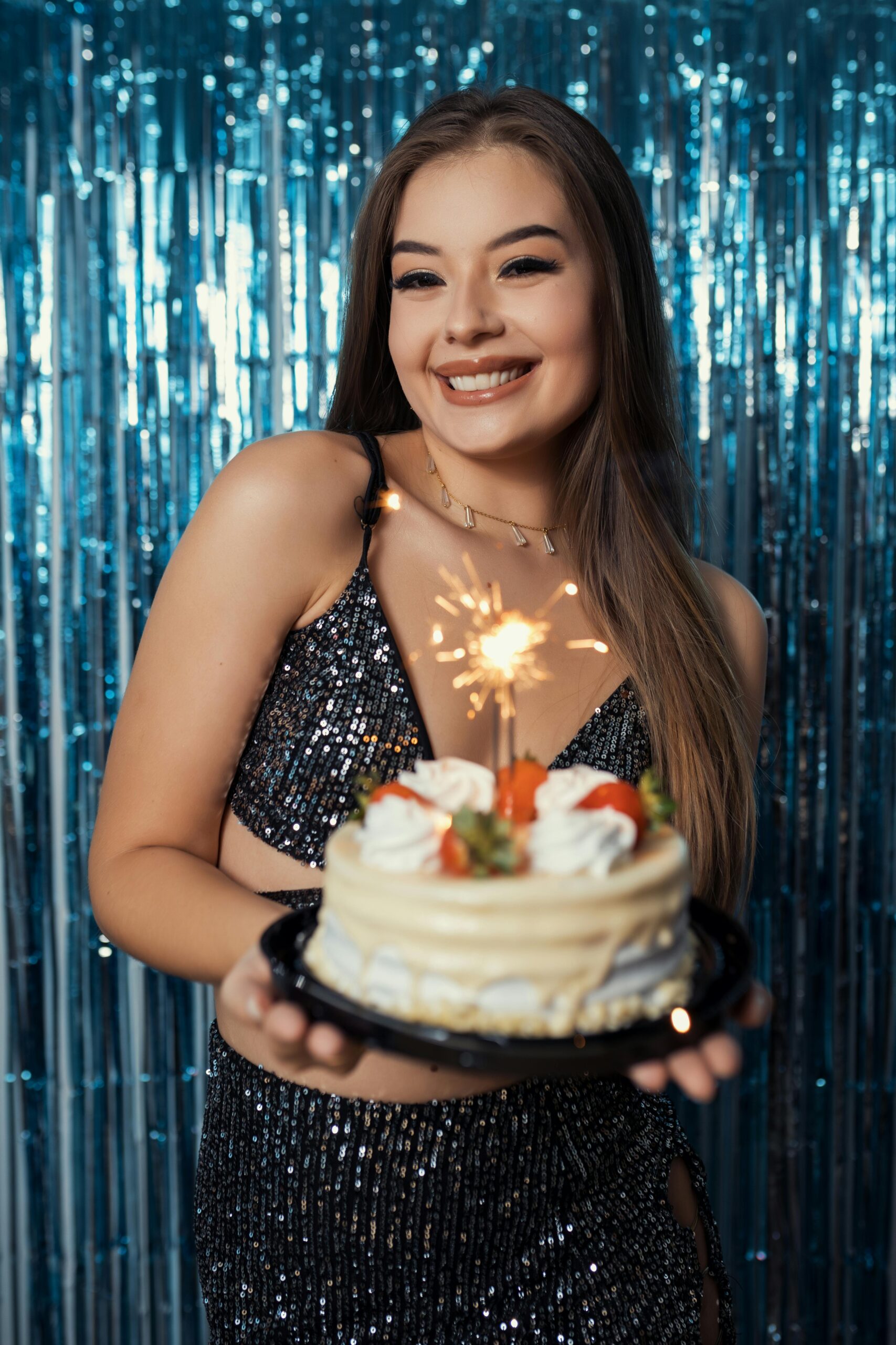 Smiling young woman holding a birthday cake with sparklers in a festive indoor setting.